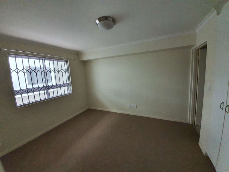 A clean, empty bedroom featuring neutral walls, carpeted flooring, built-in cupboards, and a window fitted with security bars.