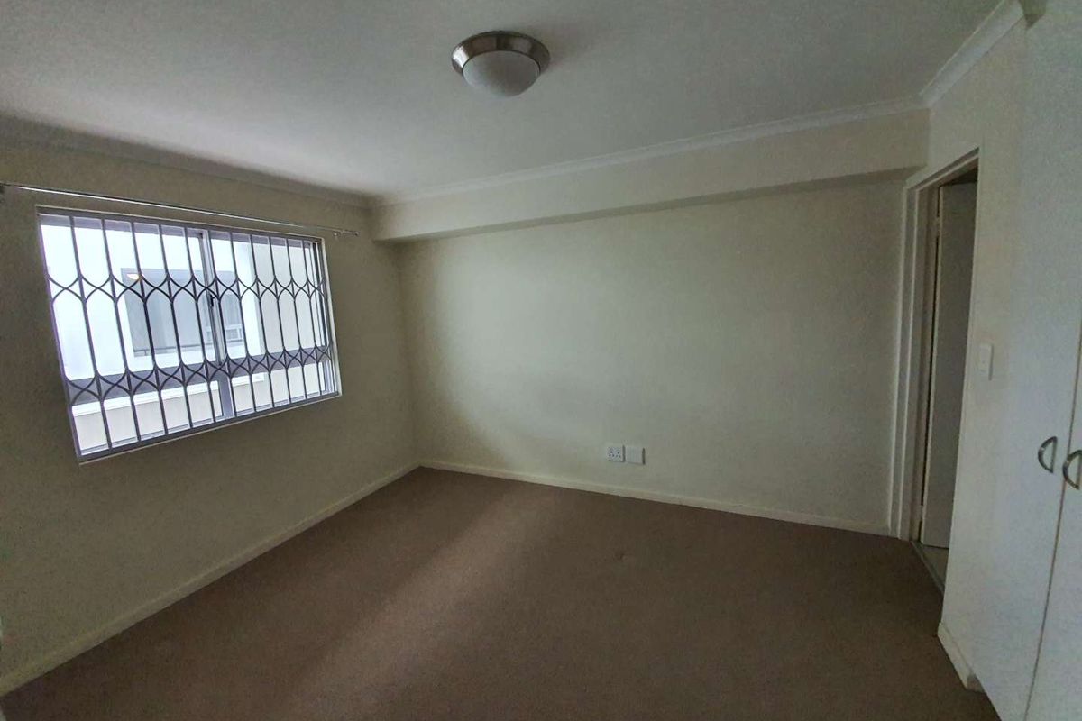 A clean, empty bedroom featuring neutral walls, carpeted flooring, built-in cupboards, and a window fitted with security bars.