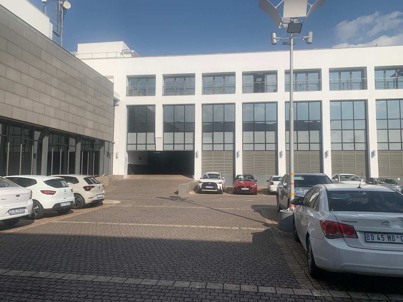 A modern multi-story office building exterior featuring a paved parking area and large glass facade windows.