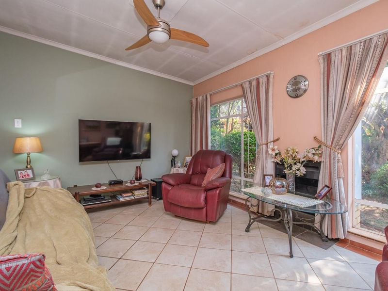 A living room featuring tiled flooring, a ceiling fan, and a fireplace, with large windows providing natural light and garden views.
