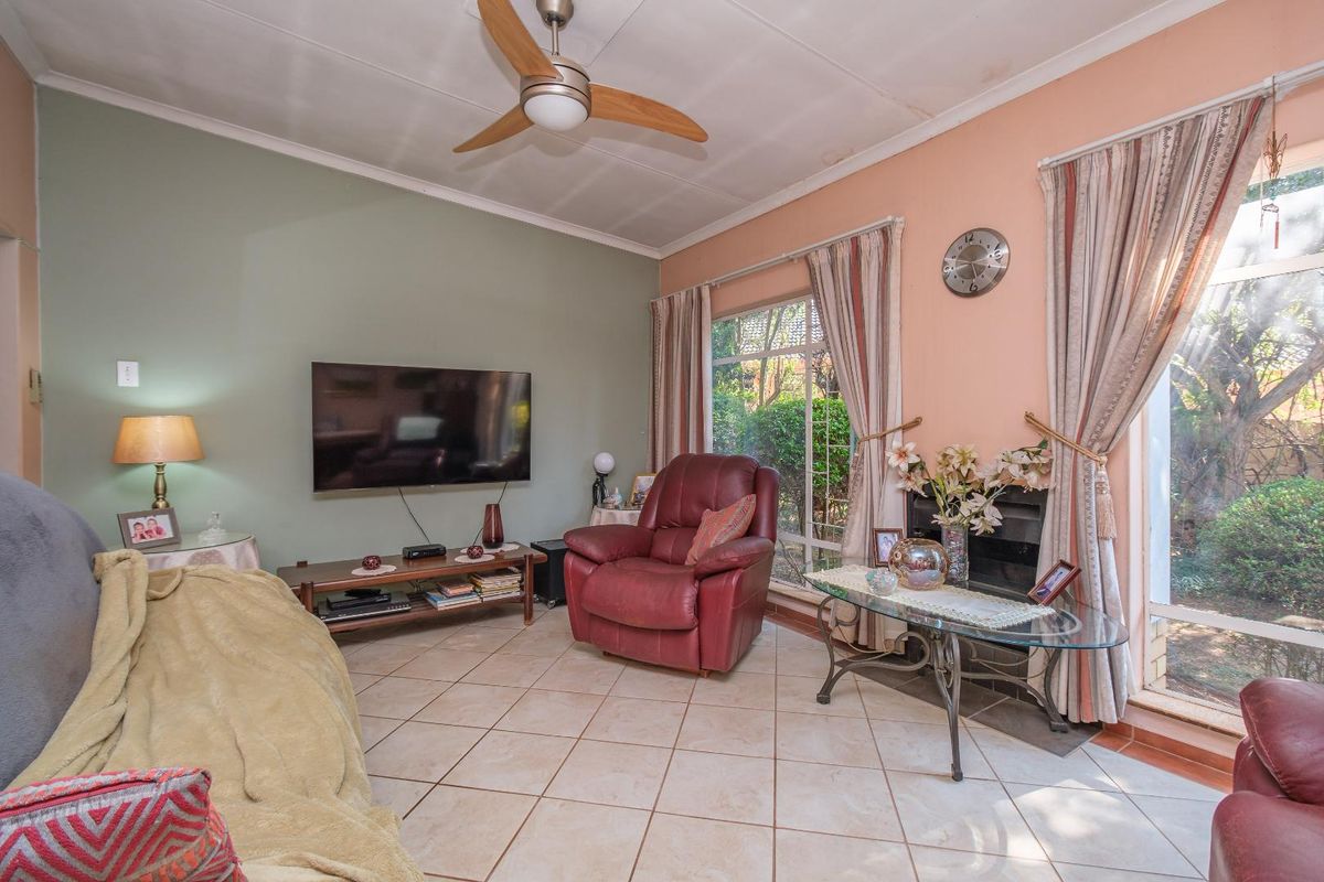 A living room featuring tiled flooring, a ceiling fan, and a fireplace, with large windows providing natural light and garden views.