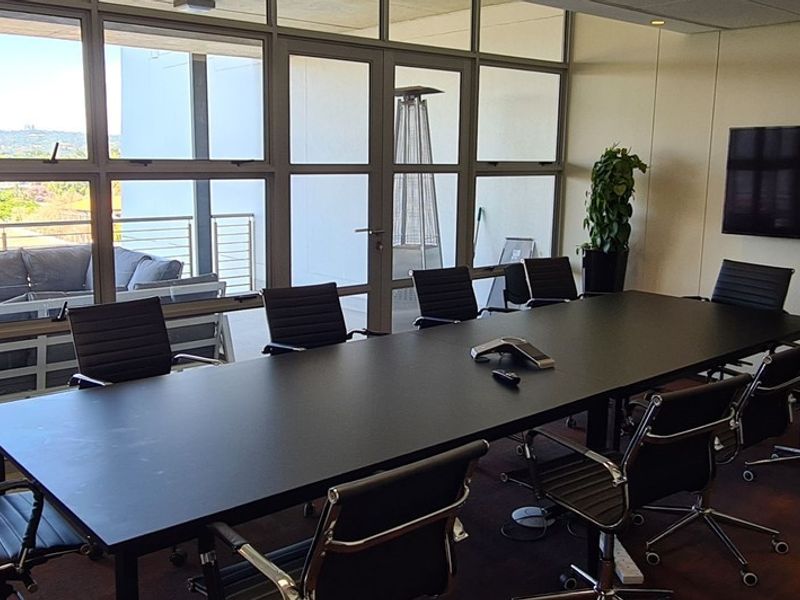 A professional boardroom featuring a large conference table, wall-mounted screen, and floor-to-ceiling glass partitions providing access to an outdoor balcony area.
