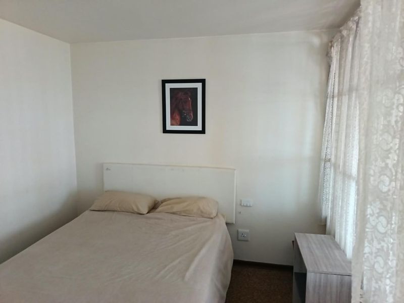 A simple bedroom featuring neutral walls, carpeted flooring, and a window with lace curtains.