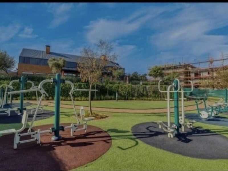 Outdoor fitness area featuring fixed exercise equipment set on rubberized safety flooring within a landscaped garden setting.