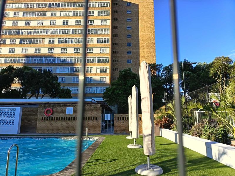 Communal swimming pool area featuring artificial turf, brick walling, and a metal security fence, set against a multi-story apartment building.