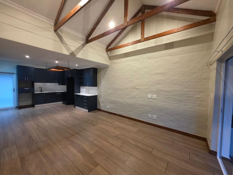 Spacious open-plan living area featuring high vaulted ceilings with exposed timber trusses, modern dark cabinetry, and wood-look tile flooring throughout.