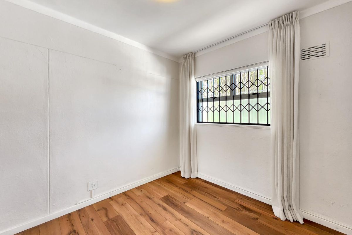A bright, empty bedroom featuring warm-toned laminate flooring, a window with a security gate, and neutral white walls.