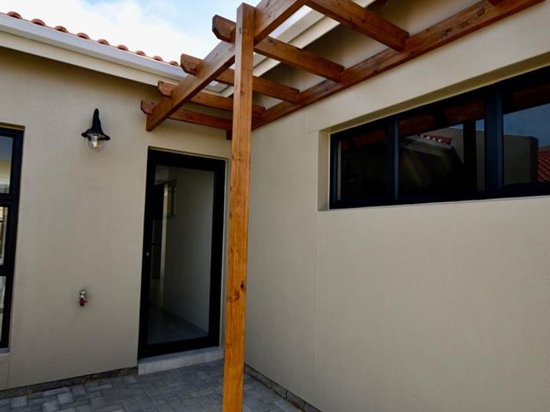 A clean, paved patio area featuring a timber pergola, modern aluminium-framed windows and door, and exterior wall lighting.