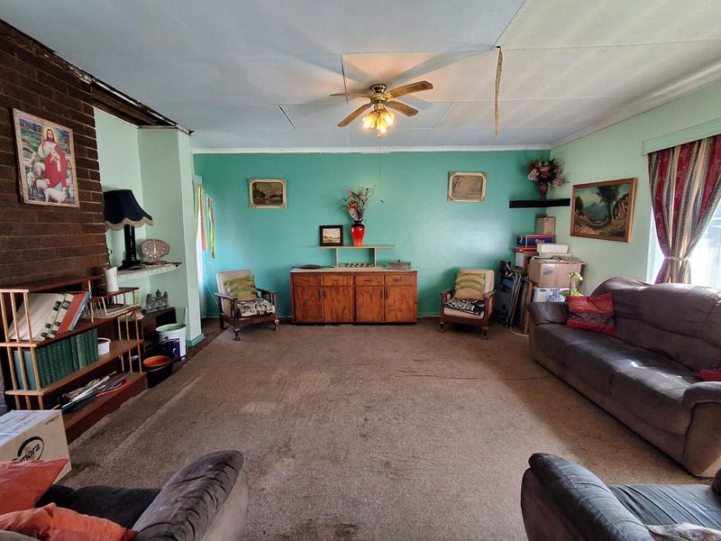 Spacious living room featuring a brick fireplace, ceiling fan, and carpet flooring, currently requiring cosmetic updates and ceiling repairs.