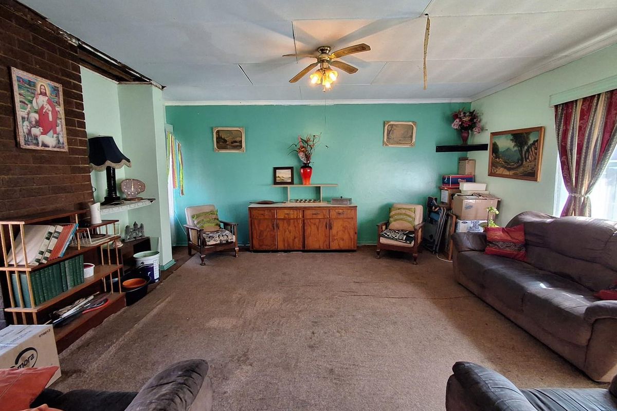 Spacious living room featuring a brick fireplace, ceiling fan, and carpet flooring, currently requiring cosmetic updates and ceiling repairs.