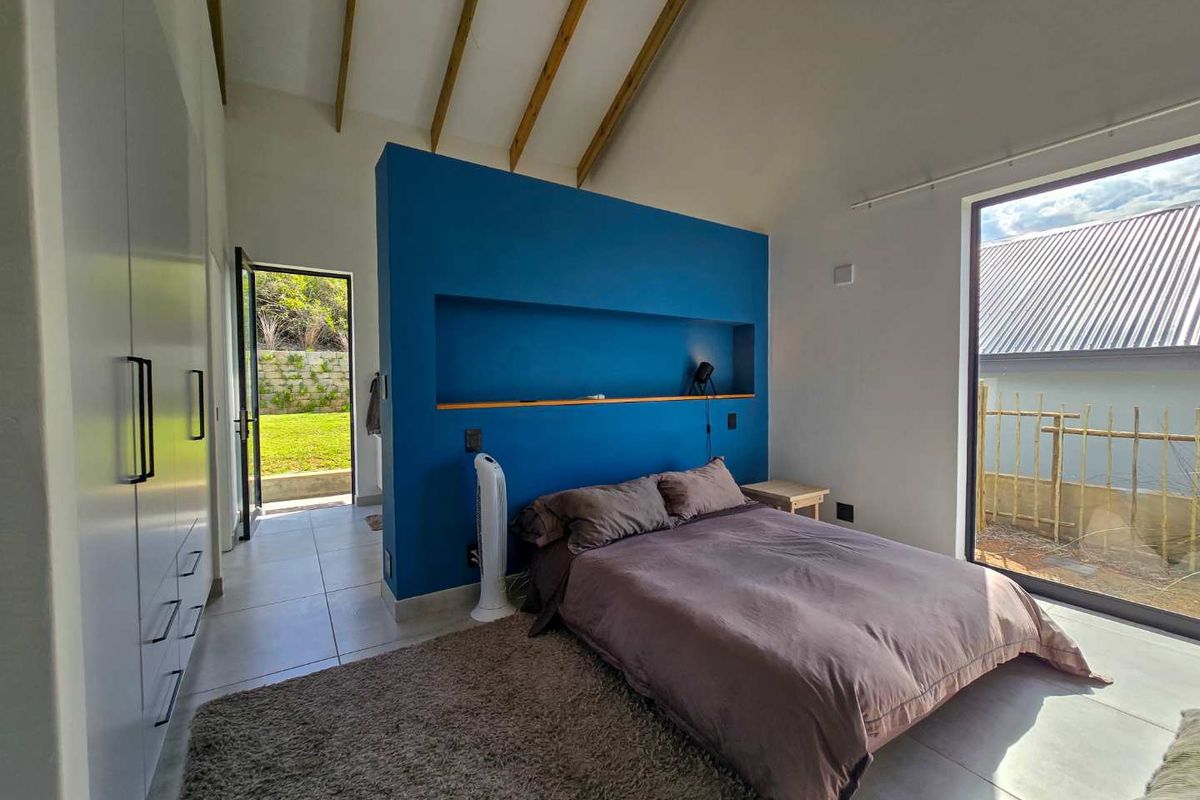 Modern bedroom featuring a vaulted ceiling with exposed timber beams, built-in wardrobes, a feature blue wall with recessed shelving, and large windows.