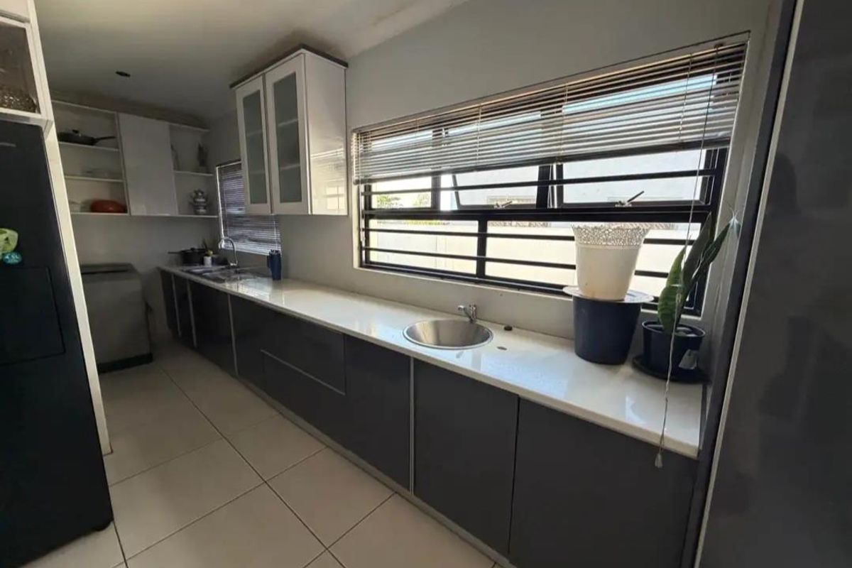 Modern galley-style kitchen featuring sleek dark cabinetry, white stone countertops, stainless steel sink, and large windows with security bars.