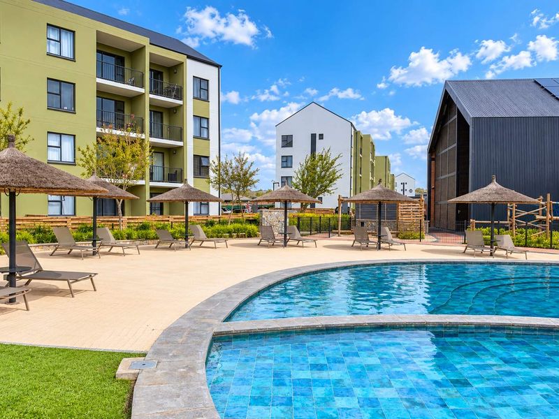 A well-maintained communal swimming pool area featuring a paved deck, thatched umbrellas, and modern apartment buildings in the background.