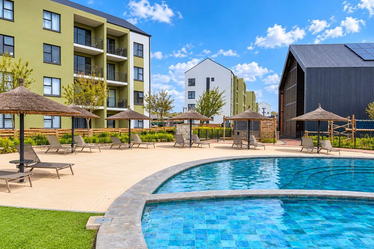 A well-maintained communal swimming pool area featuring a paved deck, thatched umbrellas, and modern apartment buildings in the background.