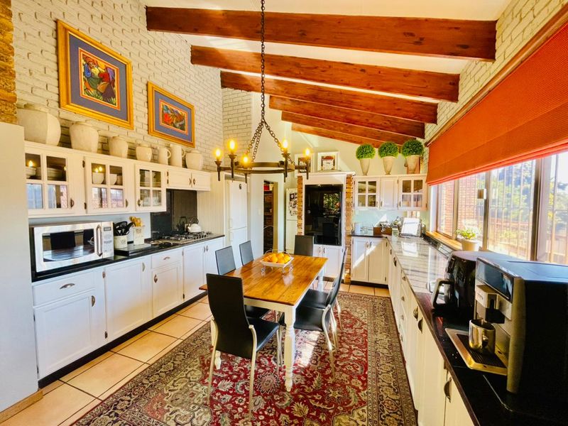 Spacious open-plan kitchen and dining area featuring exposed timber beams, painted brick walls, white cabinetry, and granite countertops with a central dining space.