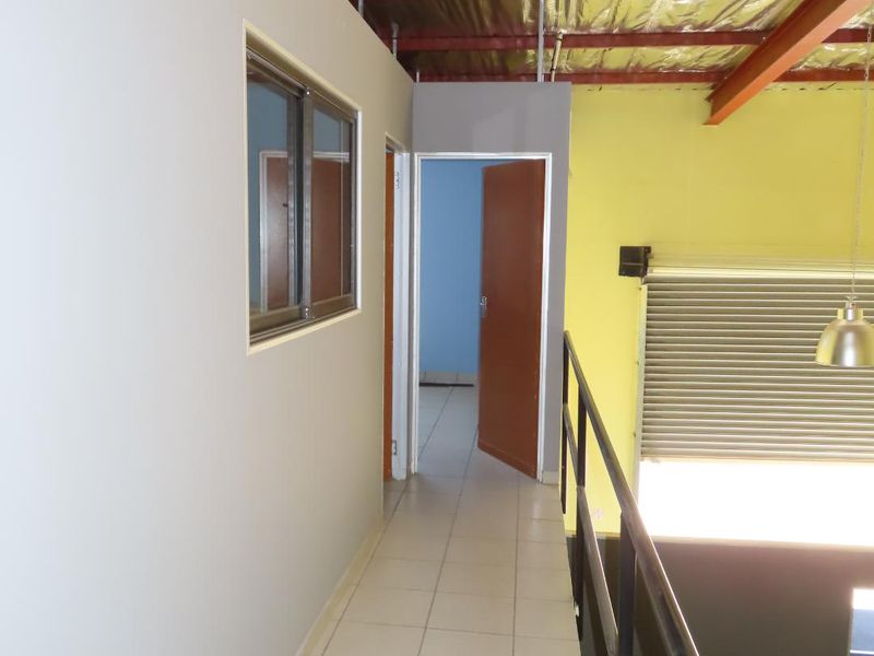 A mezzanine hallway featuring tiled flooring, metal safety railing, and an exposed industrial steel roof structure with access to an adjacent room.
