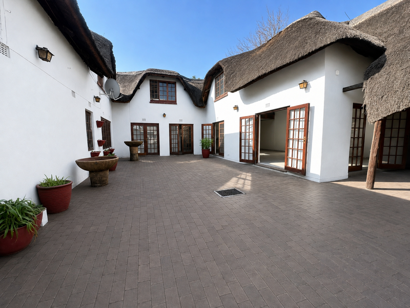 A spacious paved courtyard featuring traditional thatch roofing, multiple sets of wooden french doors, and integrated exterior lighting.