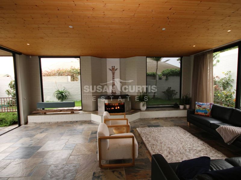 Spacious living room featuring a built-in fireplace, natural stone flooring, and a warm timber ceiling with expansive windows overlooking the garden.