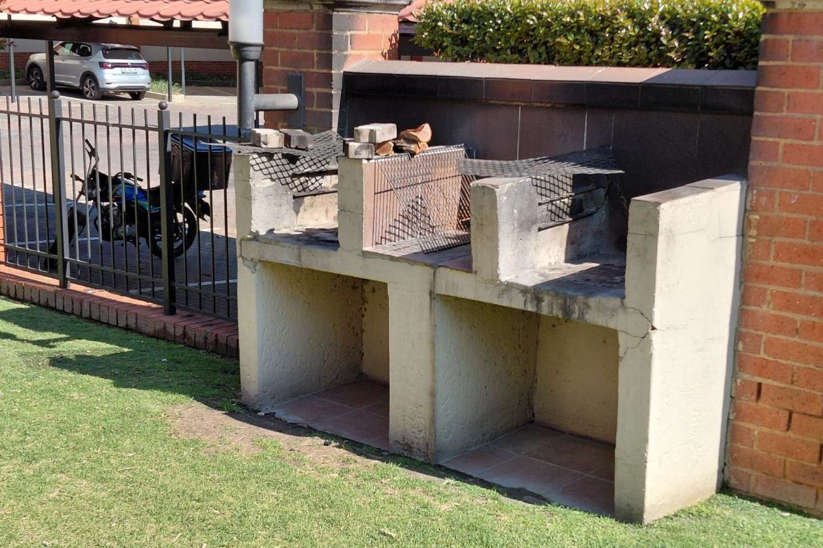 A built-in outdoor braai area featuring a concrete structure situated against a brick boundary wall in a communal garden space.