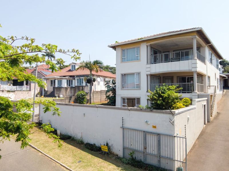 A multi-story residential building featuring balconies, a paved driveway, and a secure perimeter wall with electric fencing.