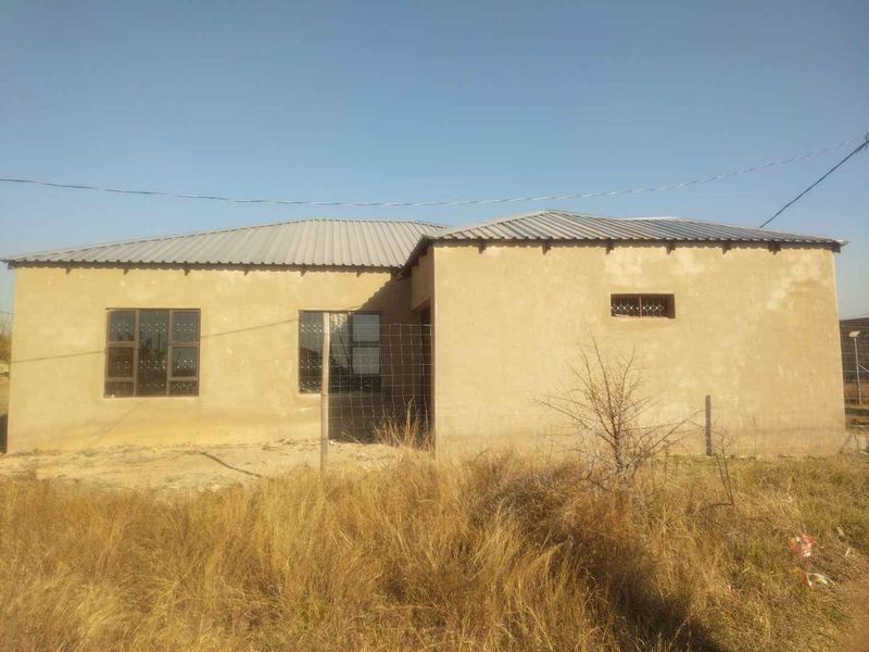 A single-story residential house featuring a metal roof and steel window frames, set on an undeveloped plot with wire fencing.