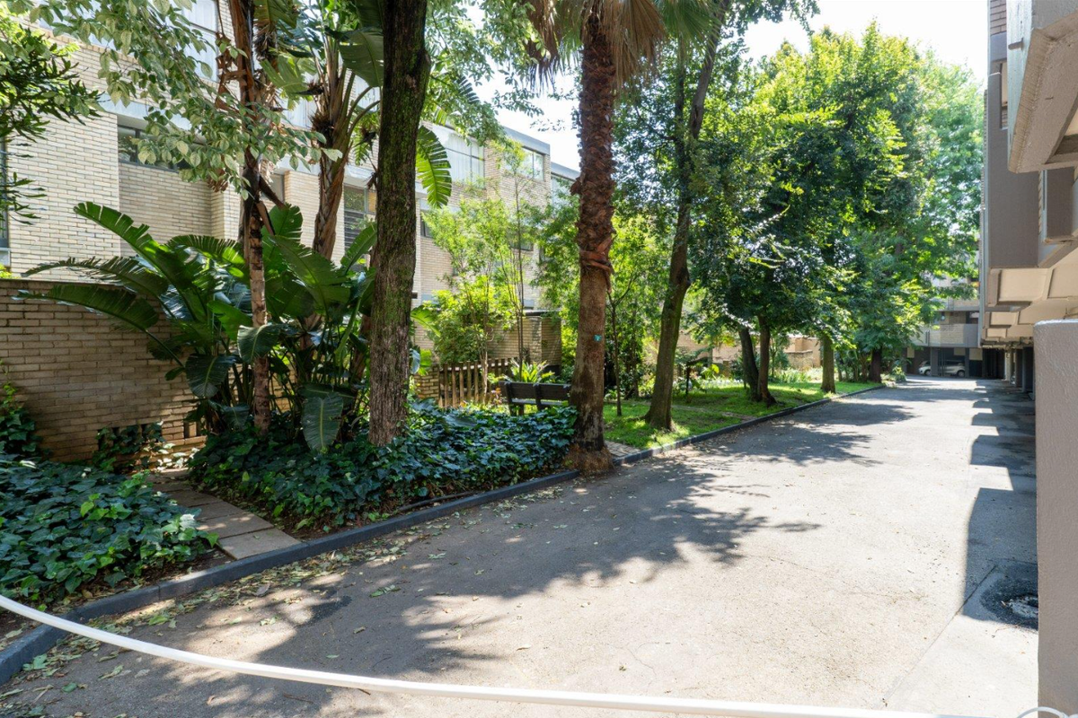A view of the apartment complex rear exterior, featuring an asphalt driveway, established trees, and landscaped garden areas alongside the brick building facade.