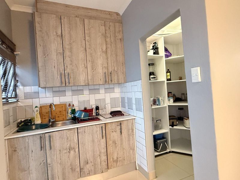 Functional scullery area featuring wood-look cabinetry, a stainless steel sink, tiled backsplash, and an integrated walk-in pantry with ample shelving.