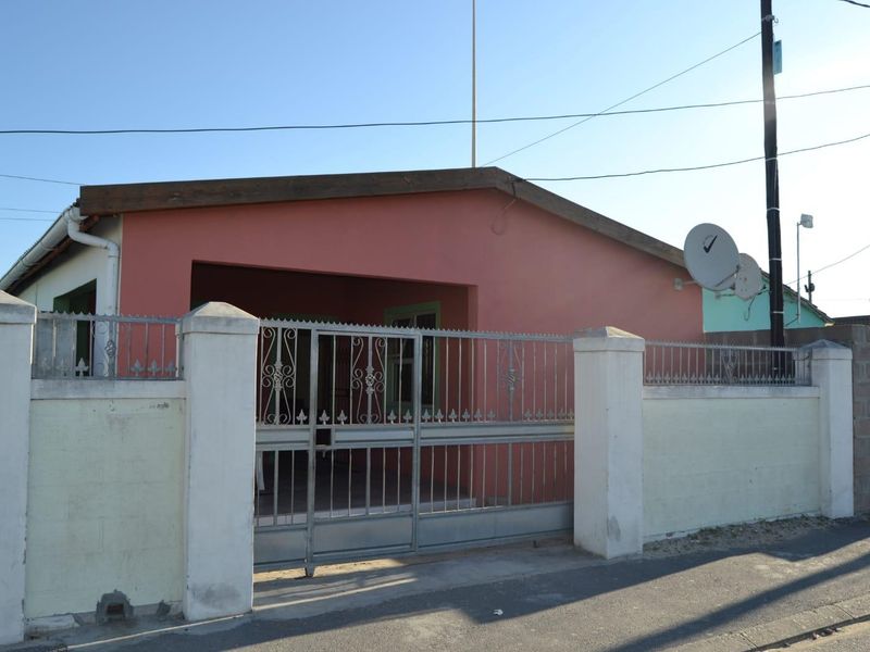 Front exterior view of a single-story house featuring a metal perimeter fence, sliding gate, and concrete boundary wall with visible signs of weathering.