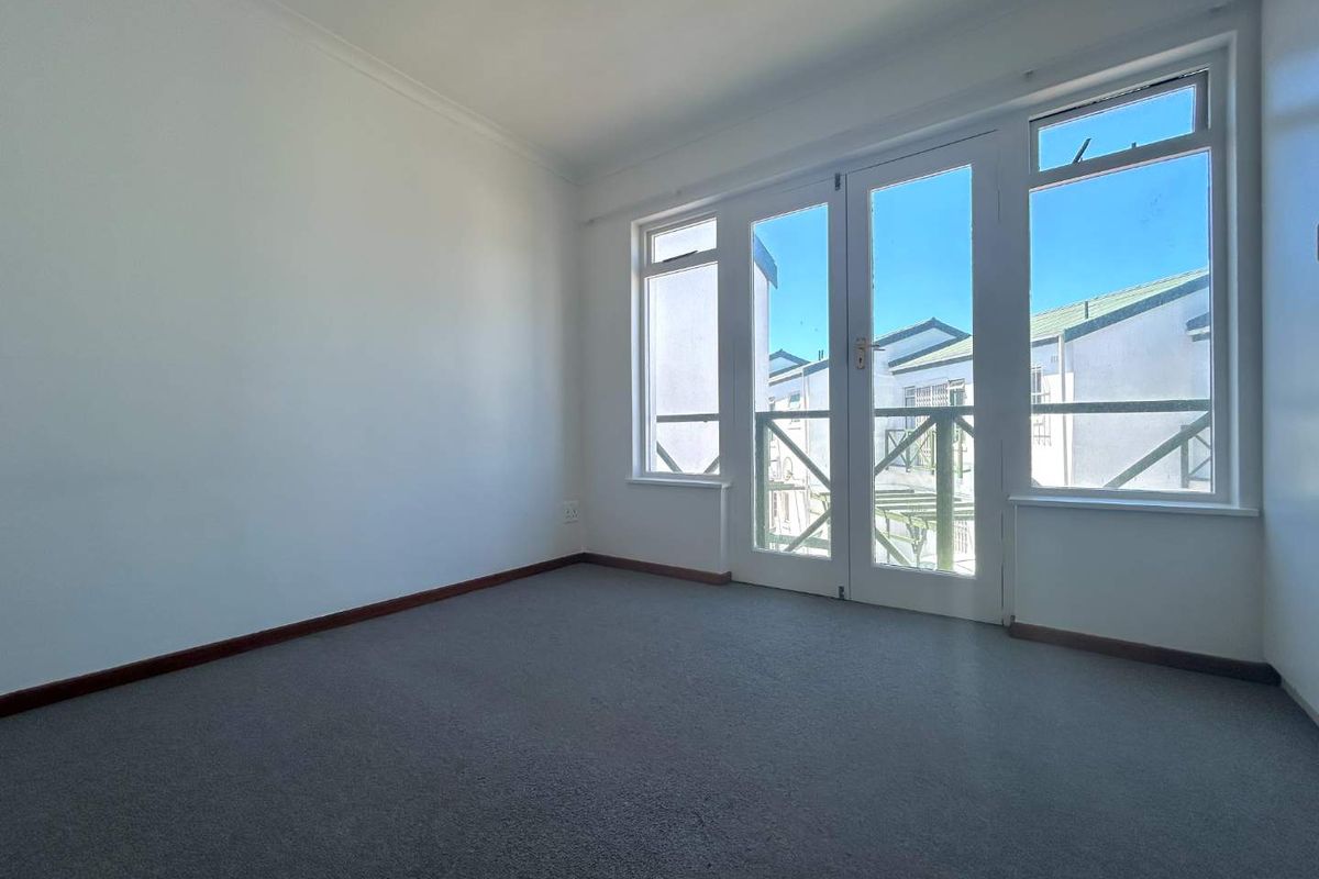 A bright, empty bedroom featuring neutral walls, grey carpeting, and glass-paned double doors leading to a balcony.