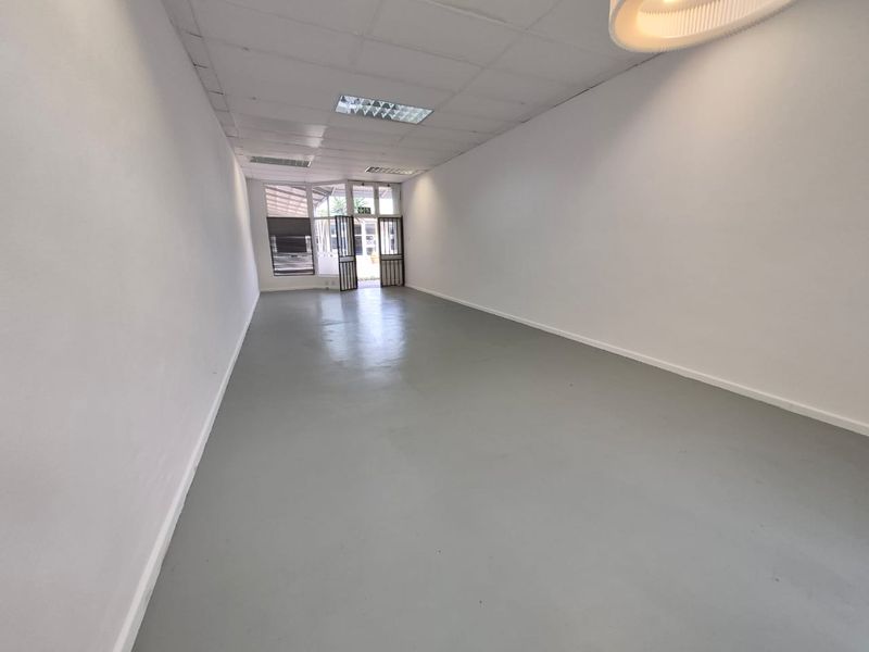 A spacious, empty retail unit featuring a clean painted concrete floor, suspended ceiling with recessed lighting, and a secure glass storefront entrance.