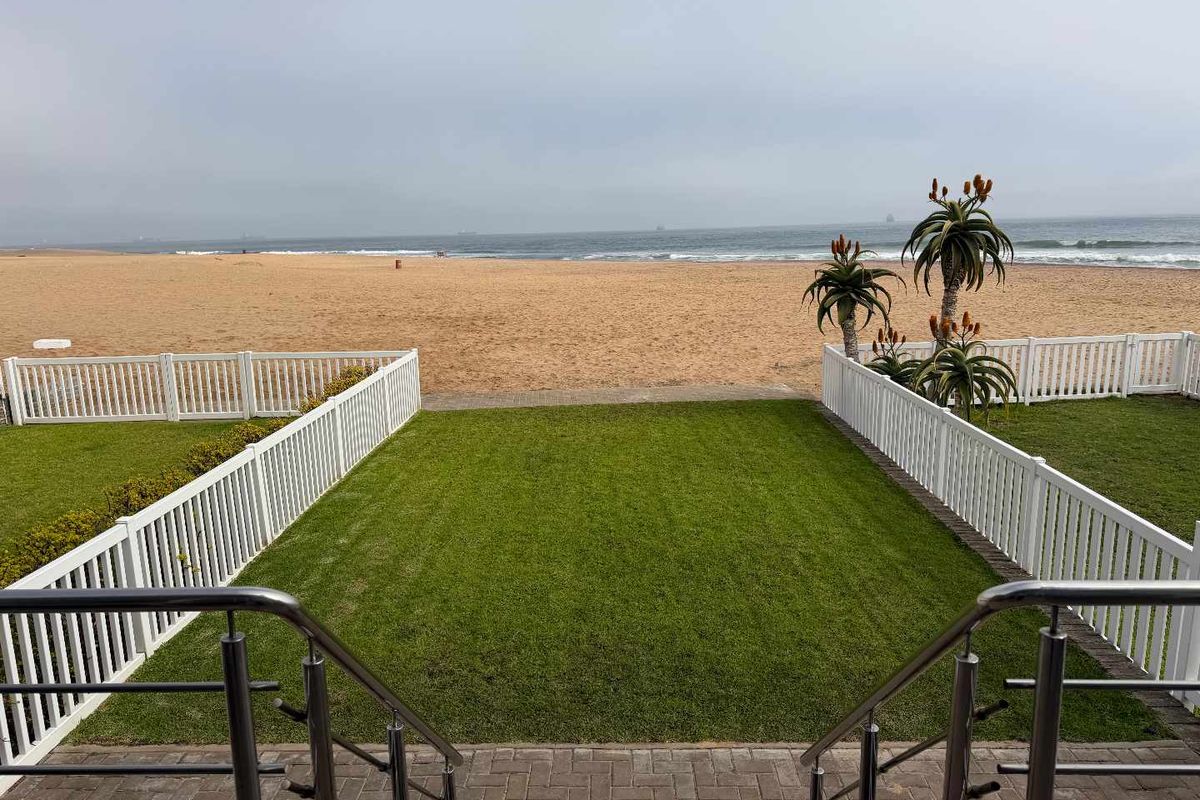 A well-maintained rear garden featuring a paved patio, stainless steel railings, and a lawn, offering direct access to the beach.