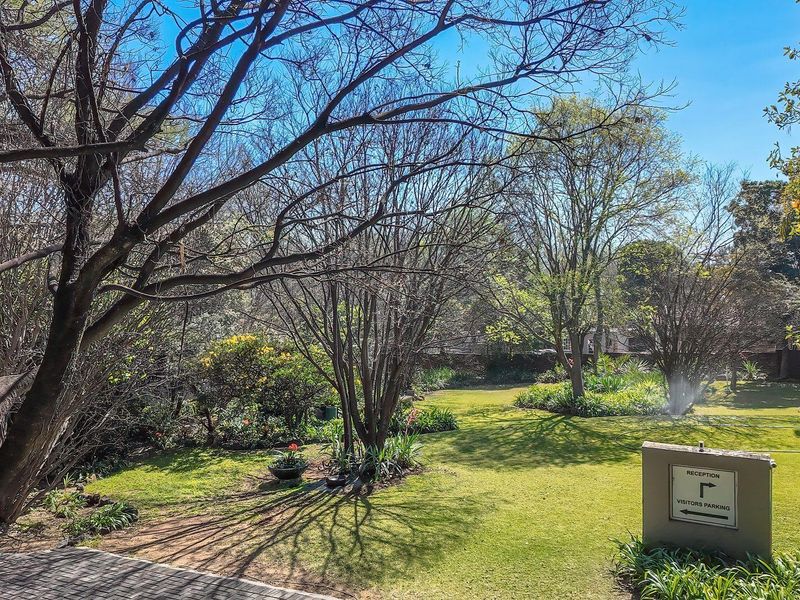 A well-maintained office garden area featuring mature trees, manicured lawn, paved pathways, and clear directional signage for visitors and parking.