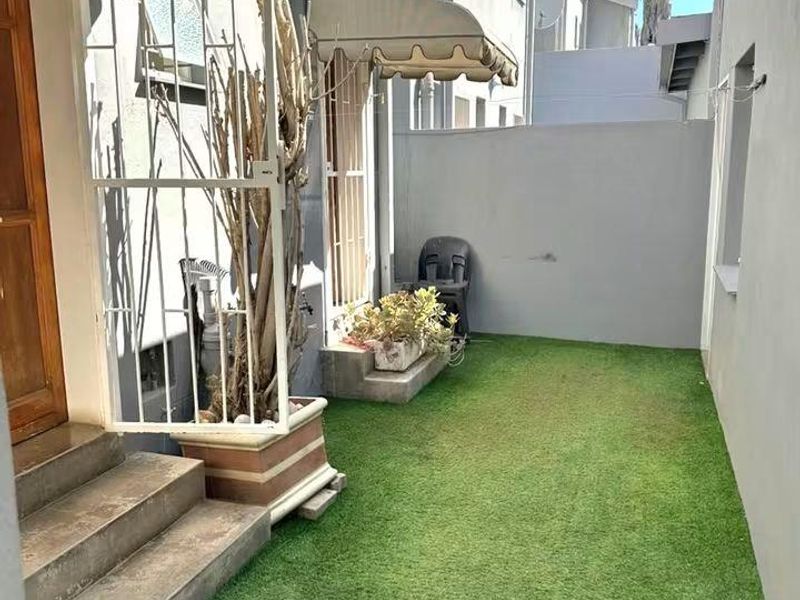 A compact rear courtyard featuring artificial turf, concrete steps, and retractable canvas awnings providing shade to the building entrance.