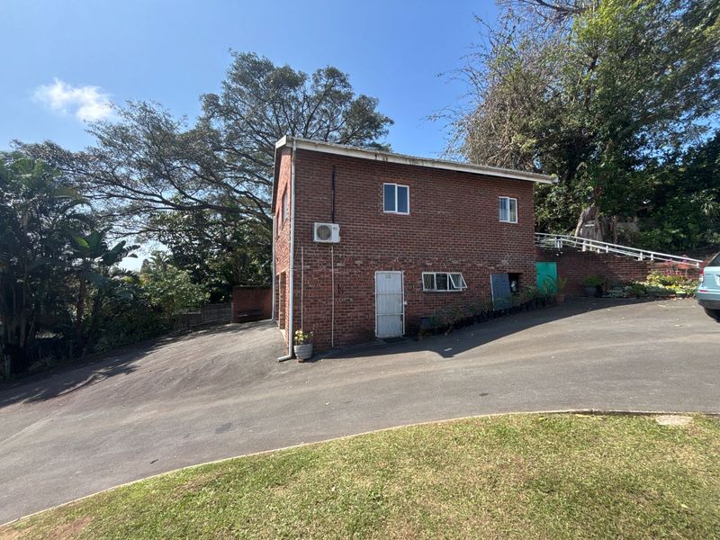 A two-story brick exterior facade featuring an asphalt driveway, security gate, and wall-mounted air conditioning unit set within a landscaped garden area.