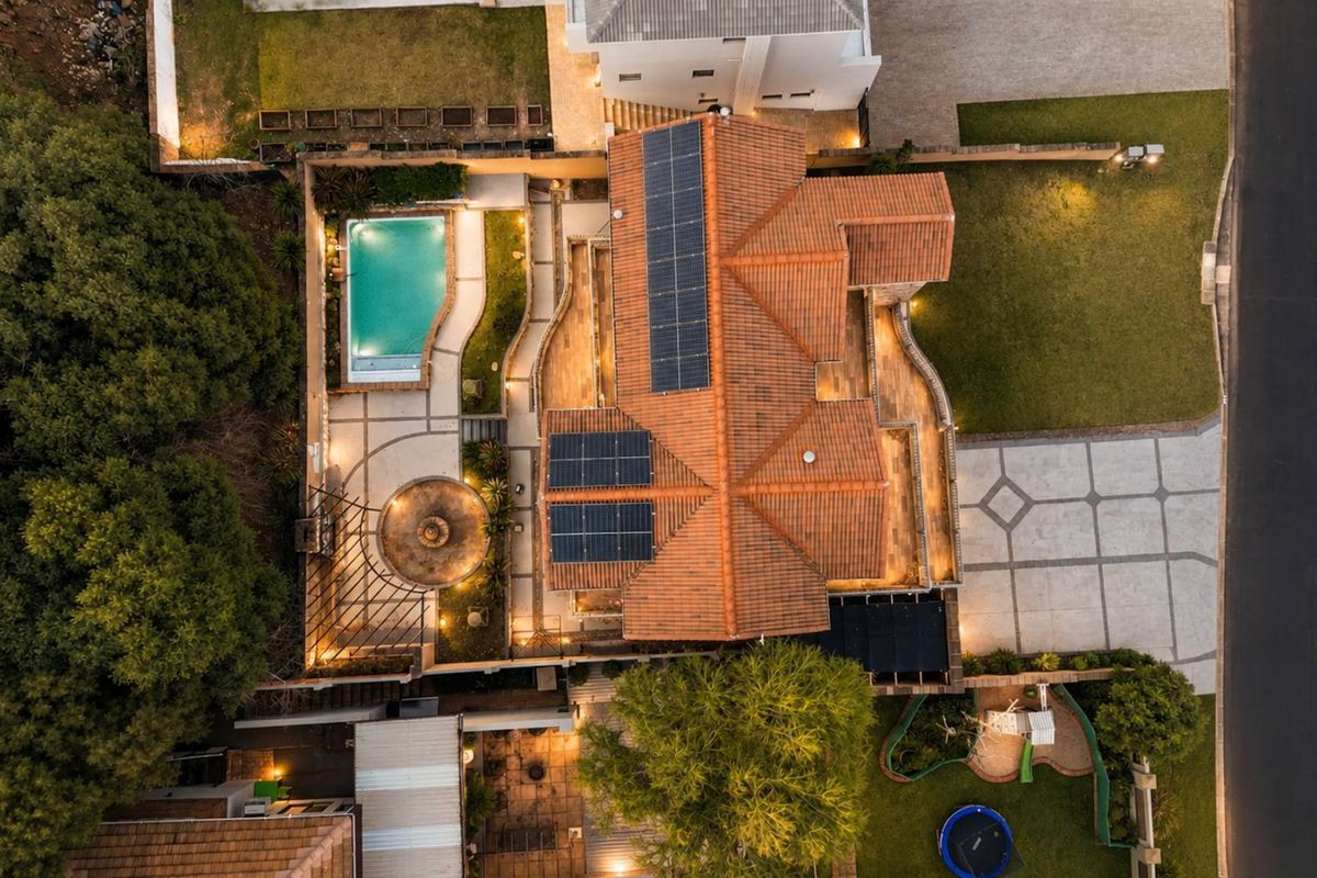 Aerial view of a residential property featuring a tiled roof with solar panels, a swimming pool, paved outdoor areas, and a landscaped garden.
