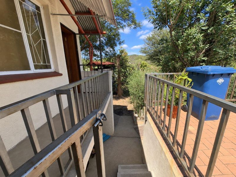 An outdoor balcony and patio area featuring metal safety railings, concrete stairs, and a corrugated metal awning, overlooking a garden space.