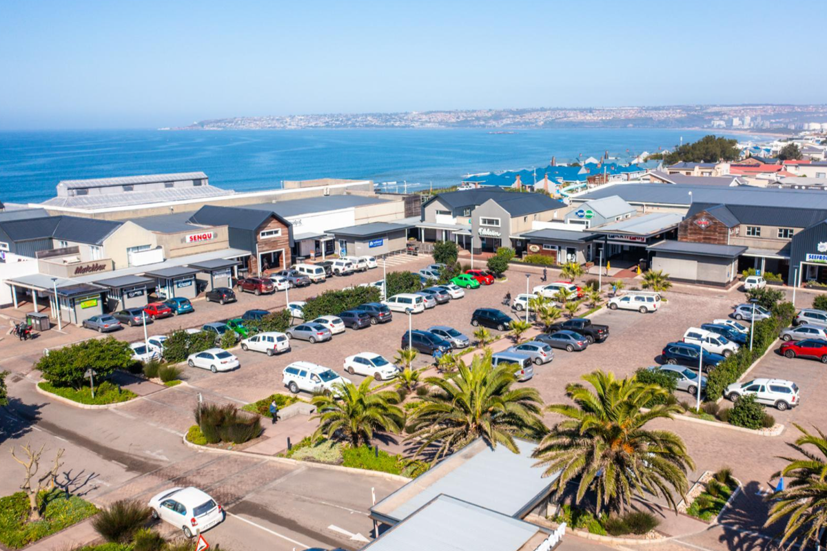 Aerial view of a commercial complex featuring multiple retail units, paved parking areas, and landscaped palm trees with a coastal backdrop.