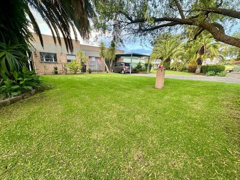 Front view of the property featuring a well-maintained lawn, paved driveway, and a carport, with established garden trees and security features on windows.