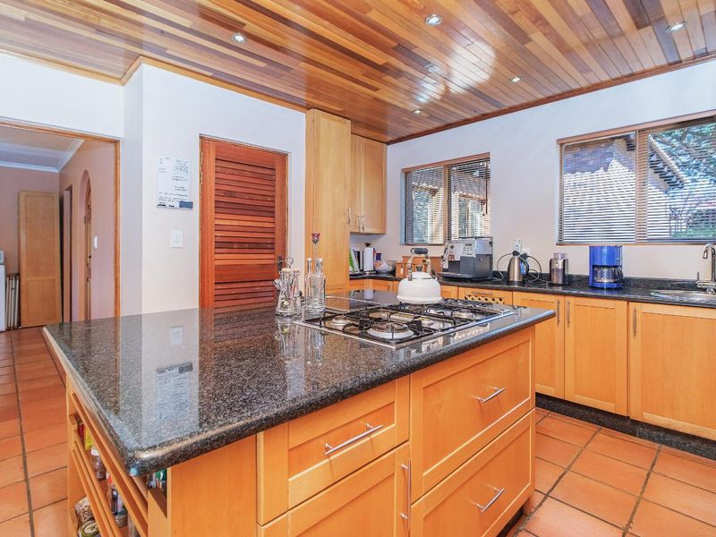 A functional kitchen featuring wooden cabinetry, granite countertops, a central island with a gas hob, and a distinctive timber-paneled ceiling.