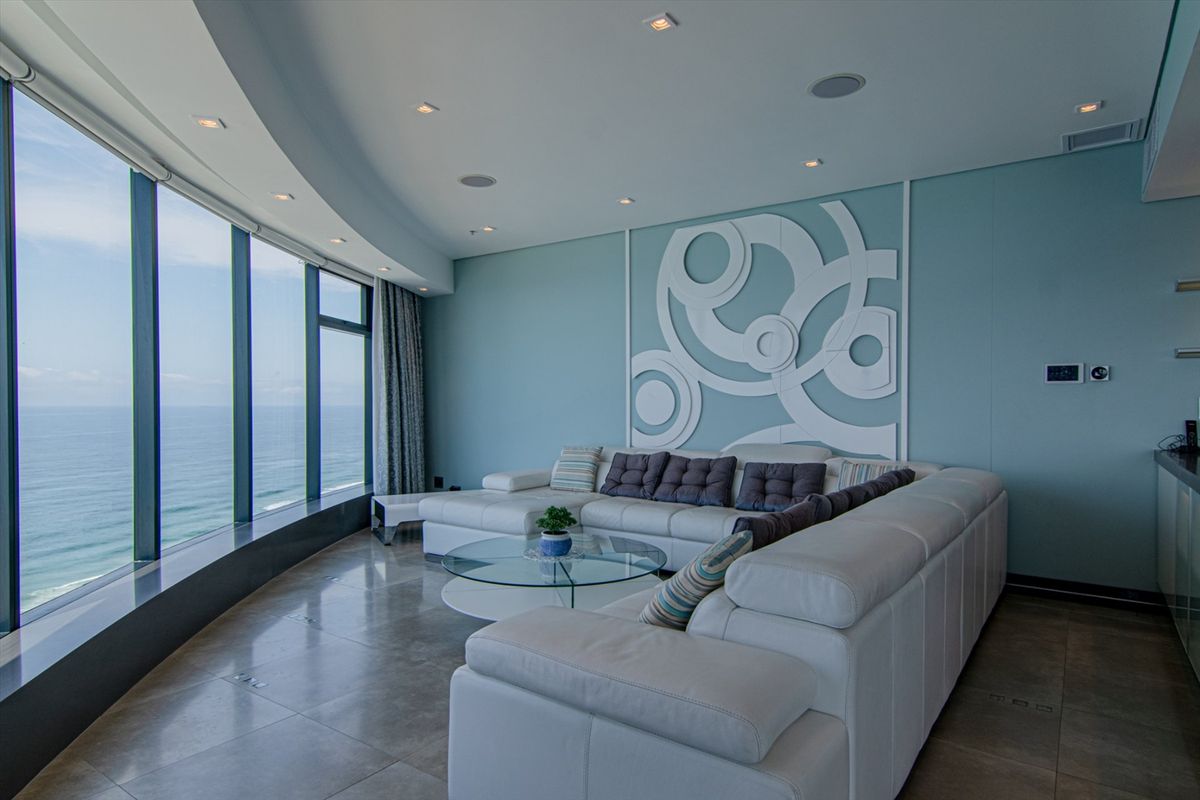 Spacious living area featuring floor-to-ceiling windows with panoramic ocean views, recessed ceiling lighting, and modern wall paneling in a contemporary penthouse setting.