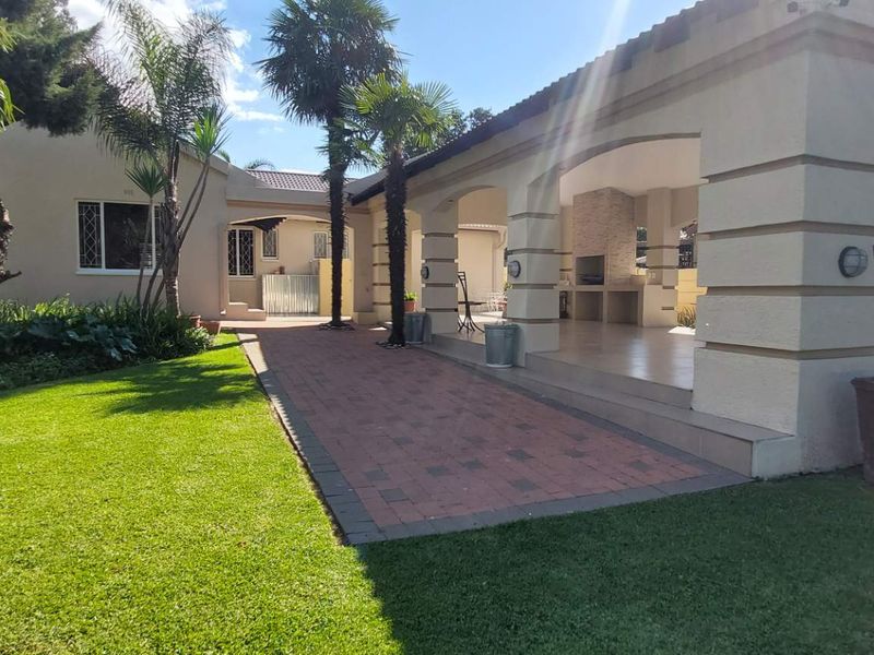 Spacious covered patio featuring a built-in braai, tiled flooring, and masonry columns, overlooking a well-maintained garden and paved walkway.