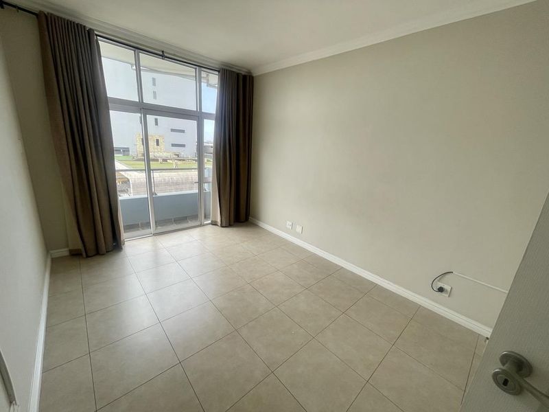 A bright, empty bedroom featuring large floor-to-ceiling windows with sliding door access to a balcony, neutral tiled flooring, and clean wall finishes.