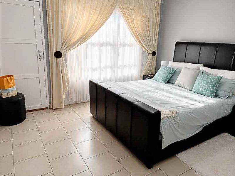 A well-maintained bedroom featuring neutral tiled flooring, light grey walls, and large windows dressed with elegant curtains.