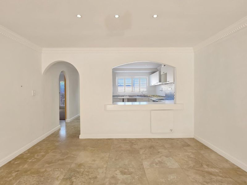 An empty living area featuring stone tile flooring, crown molding, and an arched pass-through window looking into the kitchen.