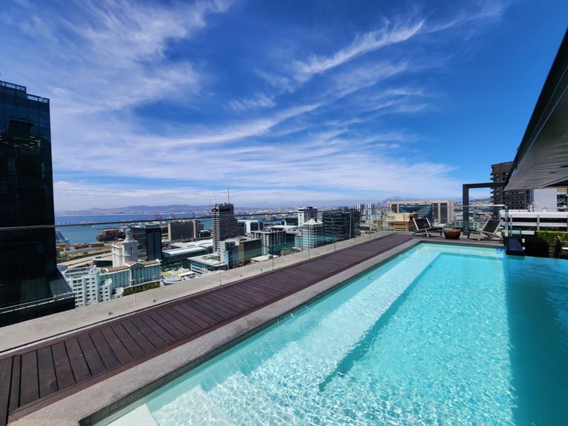 A luxury rooftop infinity pool featuring a glass balustrade and timber decking, offering expansive panoramic city and ocean views.
