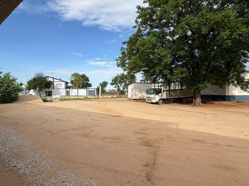 A wide view of a commercial vacant land property featuring an unpaved yard, mature trees, and existing building structures.