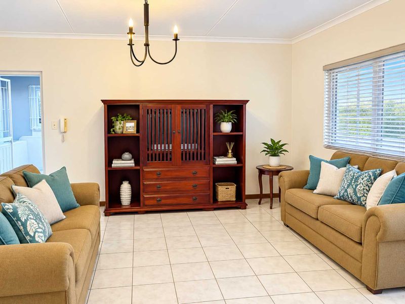 A well-maintained living room featuring neutral tiled flooring, classic chandelier lighting, and venetian blinds, offering a clean and functional space.
