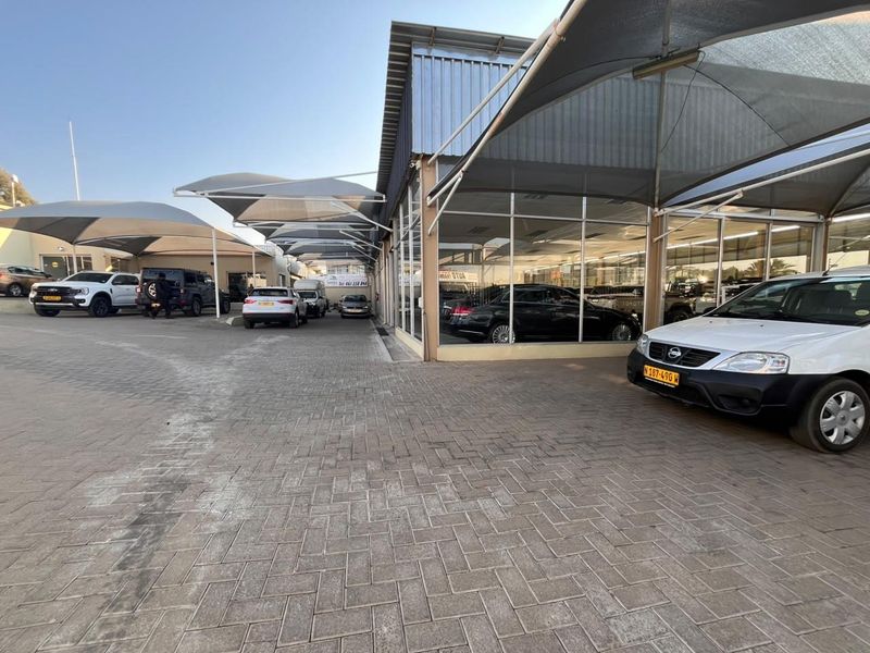 A commercial retail property featuring a paved forecourt, extensive glass display frontage, and overhead shade cloth structures for vehicle display or parking.