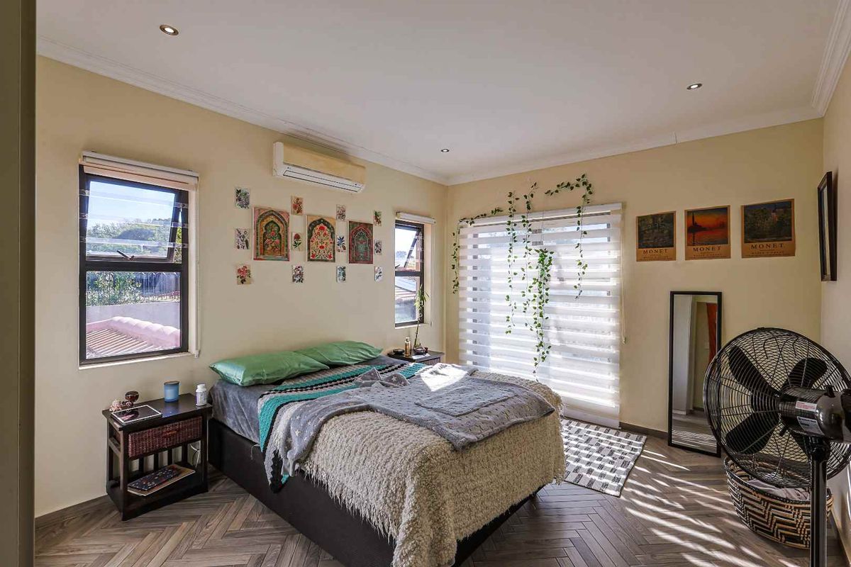 A well-lit bedroom featuring herringbone-patterned flooring, a wall-mounted air conditioning unit, and large windows with fitted blinds.