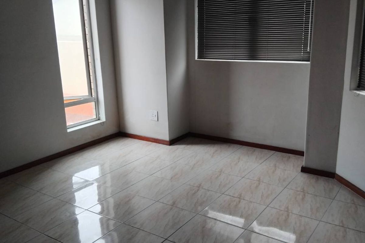 A bright, empty room featuring neutral tiled flooring, wooden skirting boards, and large windows fitted with horizontal blinds.